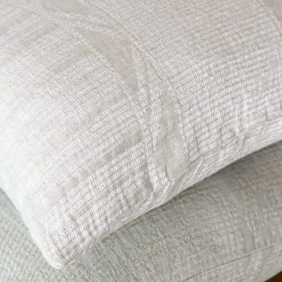 Two textured pillows on a wooden surface with a vase and branches in the background.